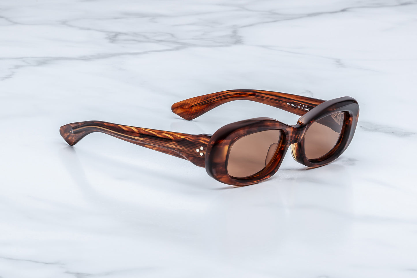 Oval sunglasses with a brown tortoiseshell acetate frame and brown tinted lenses, resting on a white marble surface.