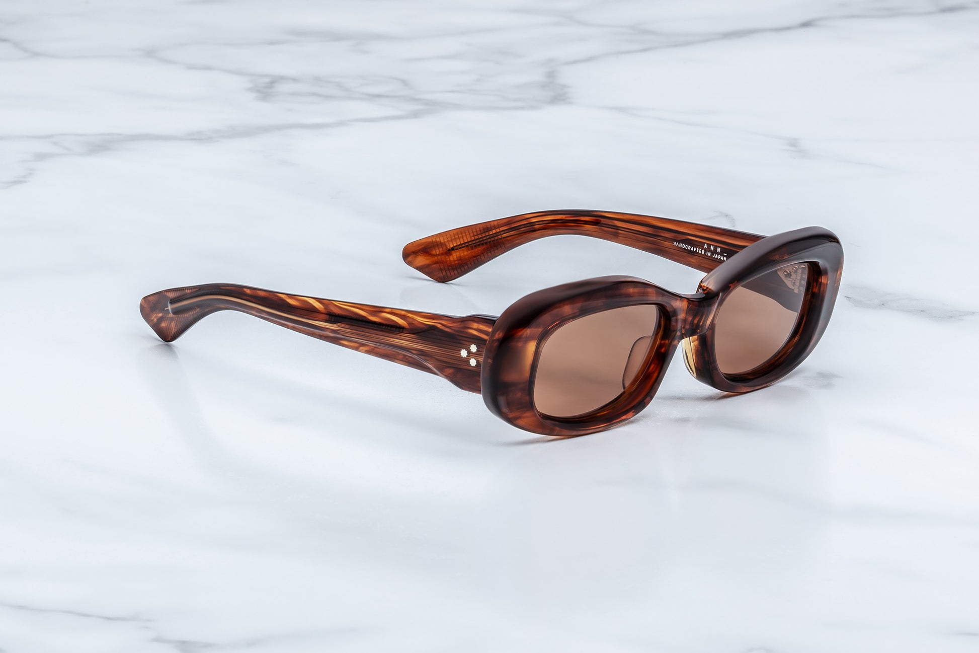 Oval sunglasses with a brown tortoiseshell acetate frame and brown tinted lenses, resting on a white marble surface.