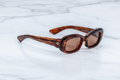 Oval sunglasses with a brown tortoiseshell acetate frame and brown tinted lenses, resting on a white marble surface.