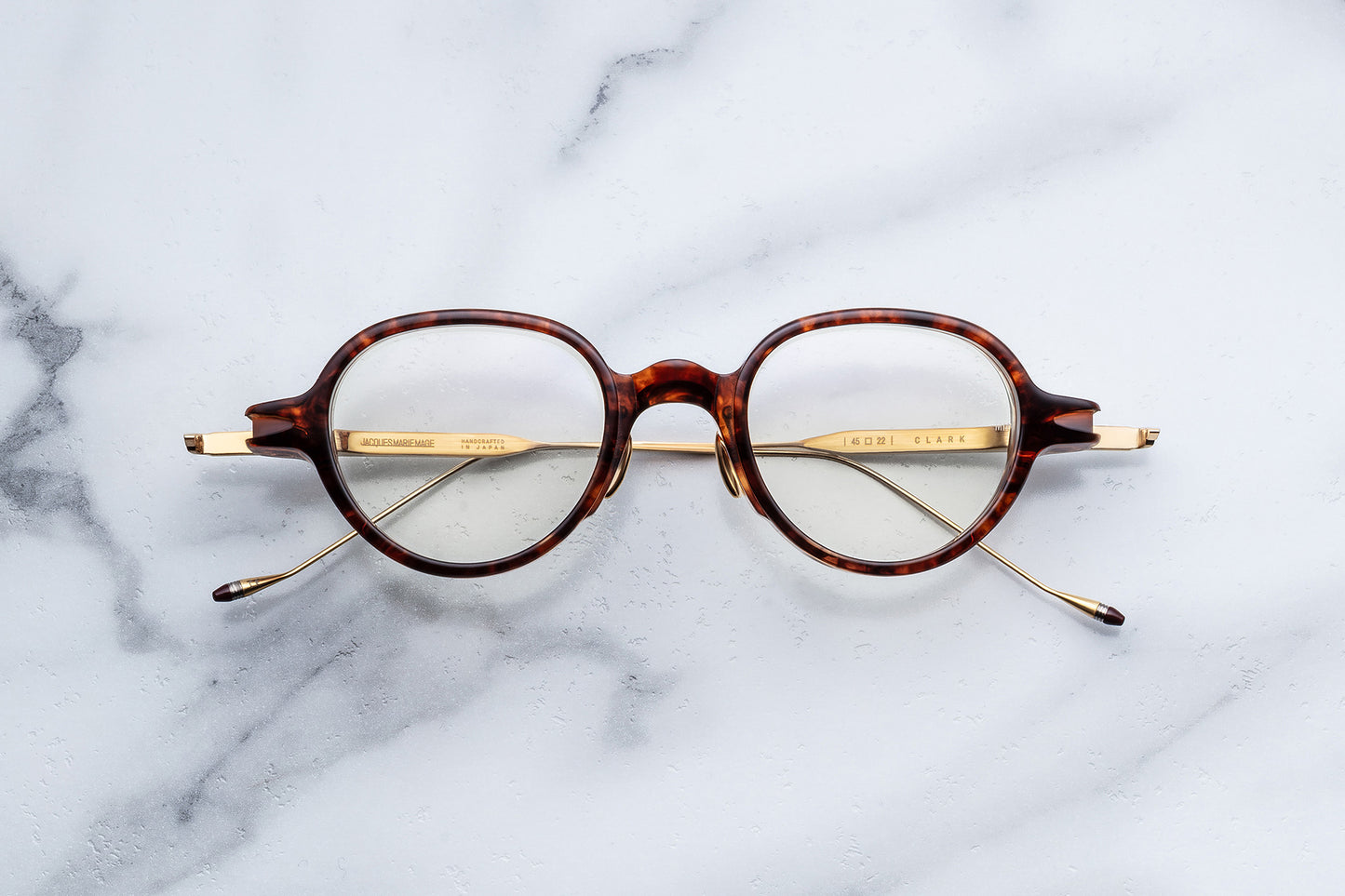 Round eyeglasses with a tortoiseshell acetate frame and gold metal temples, displayed flat on a white marble surface.