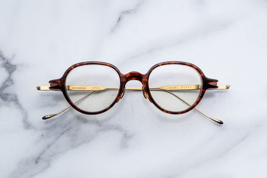 Round eyeglasses with a tortoiseshell acetate frame and gold metal temples, displayed flat on a white marble surface.