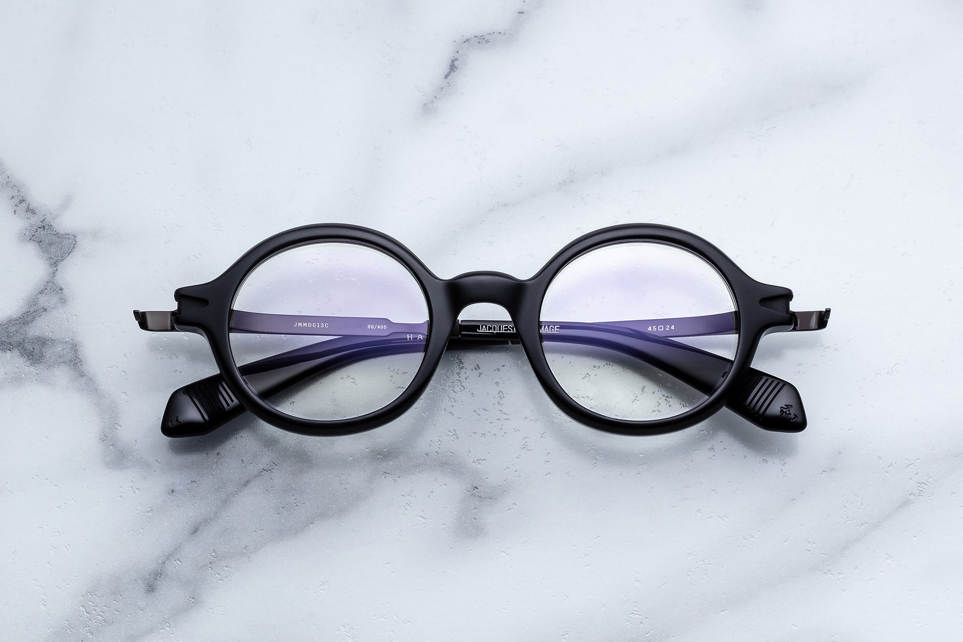 Round black eyeglass frames with purple temple arms, displayed on a white marble surface.