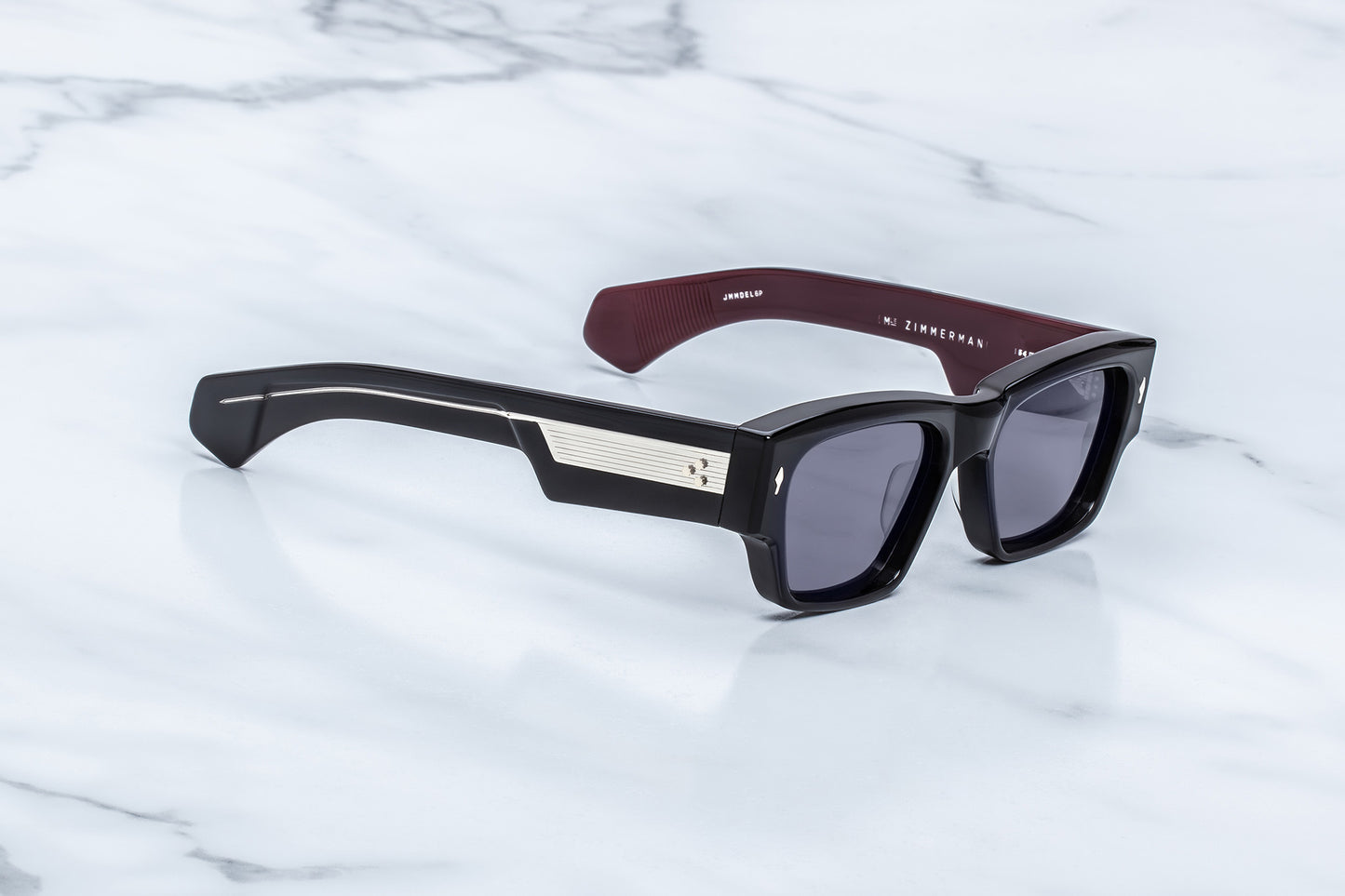 A pair of designer sunglasses with a glossy black acetate frame, a silver geometric accent on the temple, and a dark burgundy inner temple, resting on a white marble surface.