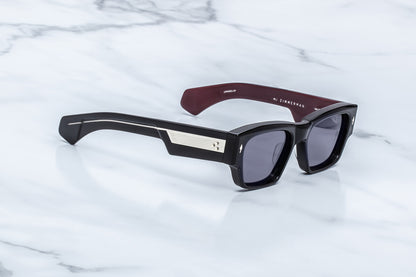 A pair of designer sunglasses with a glossy black acetate frame, a silver geometric accent on the temple, and a dark burgundy inner temple, resting on a white marble surface.